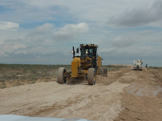 Large yellow heavy machine on a white dirt road in front of an 18 wheeler.