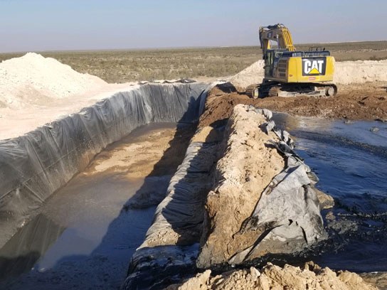Large pit construction semi full of water with yellow heavy equipment in the background.